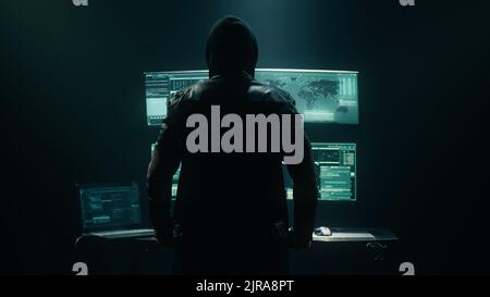 Back view of unrecognizable man in hoodie standing near desk and reading stolen data from computer, monitors in dark room before massive cyber attack on servers. Stock Photo