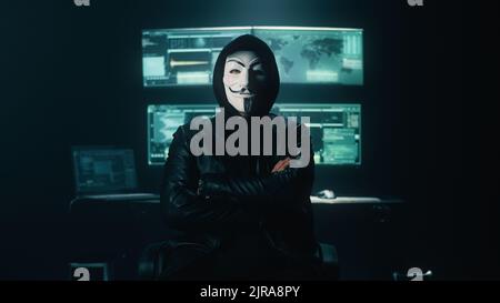 Self assured man hacker in anonymous mask crossing arms and looking at camera while standing near computer monitors in dark ambient room Stock Photo