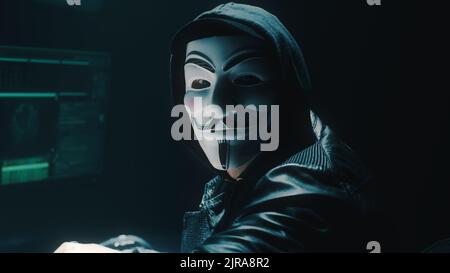 Man in Anonymous mask performing hacker cyber attack on on corporate servers and computer and looking at camera while sitting at desk in darkness Stock Photo