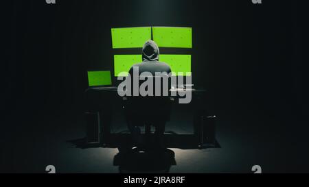 Concept shot of anonymous man in hoodie writing code to hack database while sitting at desk with computer monitors, with green screens chromakey in dim room of hacker base Stock Photo