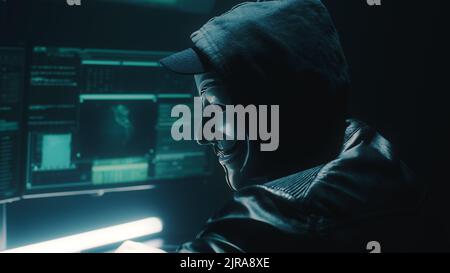A masked hacker weared black hoodie uses a computer to stage a massive data attack on big data corporate servers, while sitting at desk in dark room and hacking database Stock Photo