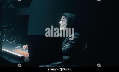 Unrecognizable fraud in mask using computer to steal data in dark room at night Stock Photo