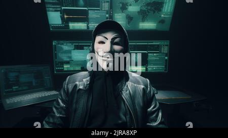 Self assured man hacker in anonymous mask crossing arms and looking at camera while standing near computer monitors in dark ambient room Stock Photo
