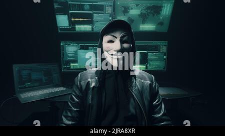 Self assured man hacker in anonymous mask crossing arms and looking at camera while standing near computer monitors in dark ambient room Stock Photo