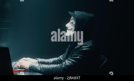 View of unrecognizable fraud in mask using computer to steal data in dark room at night Stock Photo