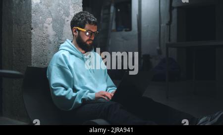 Male IT developer working with software and website at night on a laptop during the war in a dark bomb shelter Stock Photo