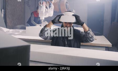 Male worker in modern VR glasses comparing plastic block with virtual plan while working in professional workshop with female colleague using robotic arm milling machine Stock Photo
