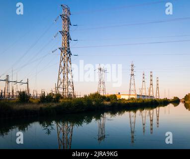 A row of metallic transmission towers of various shapes reflecting in the still water of a canal against blue sky at dusk. Stock Photo