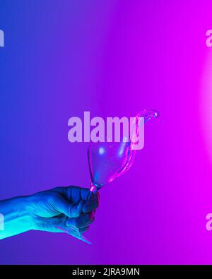 Greeting toast. Authentic hand pouring pure liquid from wine or champagne glass over bright blue-pink gradient background in neon light. Stock Photo