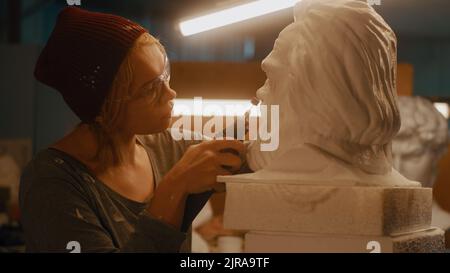 Female artisan in hat and goggles polishing bust of bearded man against lamps while working in creative studio in evening Stock Photo