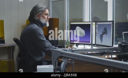 Mature man with gray hair and bread sitting at desk and creating 3D model on computer near coworker in modern design studio Stock Photo