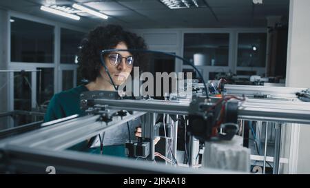 Young man in glasses with curly hair checking 3D printer with plastic model and using tablet during work in modern workshop Stock Photo