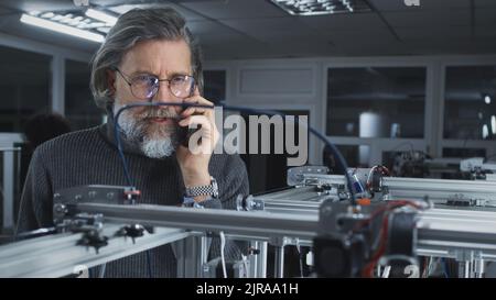 Mature bearded man in turtleneck and glasses talking with client on cellphone and watching modern printer printing 3D model in professional design studio Stock Photo