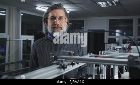 Bearded middle aged male engineer in turtleneck and glasses looking at camera while standing behind modern 3D printer printing model in professional studio Stock Photo