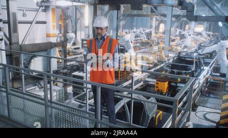 High angle of mature male engineer in uniform and hardhat using tablet to control robotic arms during work on contemporary car factory Stock Photo