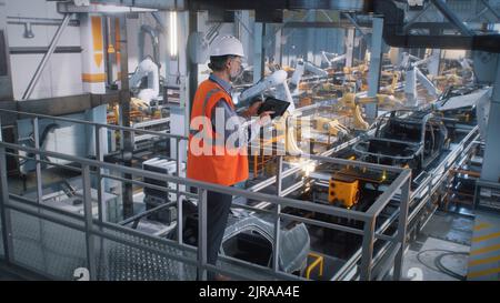 High angle of mature male engineer in uniform and hardhat using tablet to control robotic arms during work on contemporary car factory Stock Photo