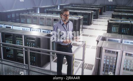 It Specialist Use Digital Pad in Big Server Farm Cloud Computing Facility or Mining center, monitoring cybersecurity and server status. High angle pan around view. Stock Photo