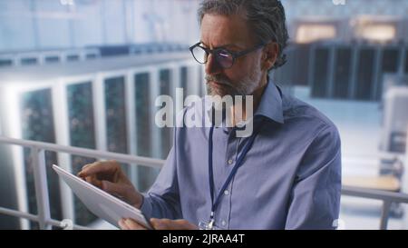 It Specialist Use Digital Pad in Big Server Farm Cloud Computing Facility or Mining center, monitoring cybersecurity and server status. High angle pan around view. Stock Photo