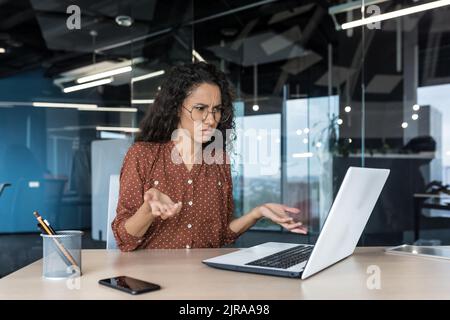 Upset and nervous business woman looking at laptop screen, female worker working inside office building, frustrated and dissatisfied with result, spreading hands Stock Photo
