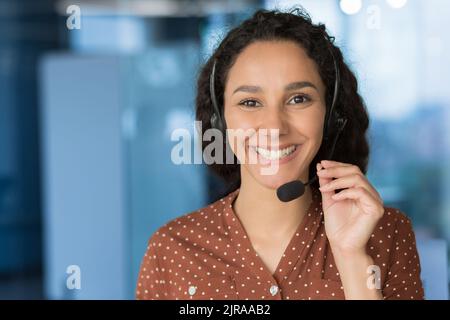 Close up photo portrait of young beautiful hispanic woman, business woman smiling and looking at camera, receptionist using headset for video call, happy and successful call center worker Stock Photo