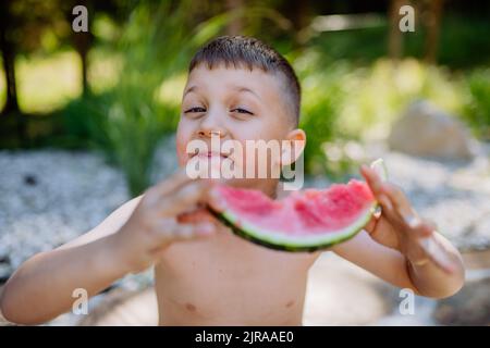 Little boy sitting near lake and eating watermelon on hot sunny day during summer vacation. Stock Photo