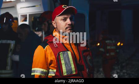 Male paramedic in uniform and hat looking at camera while standing near ambulance car during rescue mission after disaster at night Stock Photo