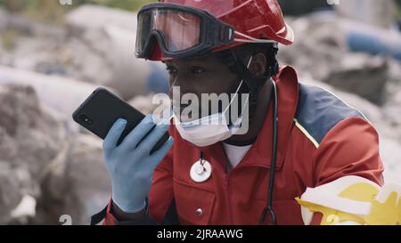 Black man in red uniform recording audio message on cellphone while sitting on rubble of demolished building after earthquake Stock Photo