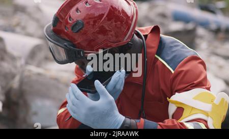 Black man in red uniform recording audio message on cellphone while sitting on rubble of demolished building after earthquake Stock Photo