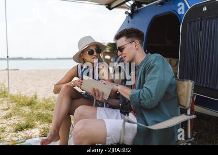 Young couple sitting together in front of van, camping and reading book. Stock Photo