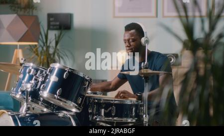 African American male musician in wireless headphones playing energetic and rhythmic tune on drums during rehearsal at home Stock Photo