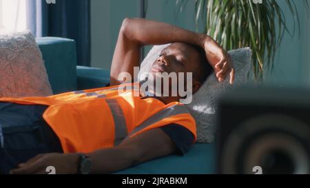 African American man in orange waistcoat yawning while lying on sofa and sleeping after work in daytime at home Stock Photo