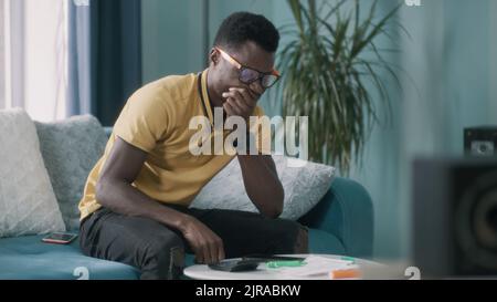 Depressed African American man in glasses looking through utility bills and thinking while sitting on couch at home Stock Photo