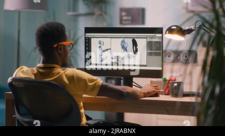 Black man in glasses and yellow t shirt sitting at table and using computer to create 3D model of futuristic robot for movie during work in home office Stock Photo