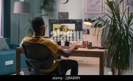 Back view of African American man retouching photo of astronaut on computer while sitting at table in workplace at home Stock Photo
