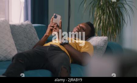 Glad African American male in casual clothes chilling on couch and using smartphone in living room in daytime at home Stock Photo
