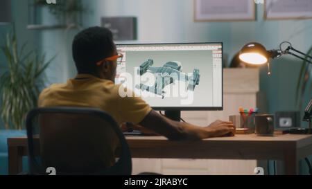 Back view of black man in yellow t shirt and glasses creating 3D model of modern aircraft on computer while sitting at table in home office and working on remote project Stock Photo
