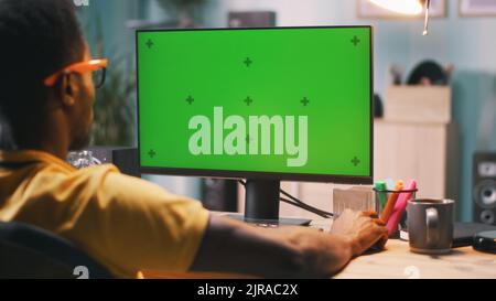 African American guy in glasses sitting at table and browsing computer with chromakey screen during work in home office Stock Photo