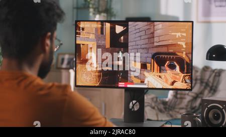 Side view of bearded Indian man in casual clothes and wireless headphones sitting at table and playing shooter on computer in weekend at home Stock Photo