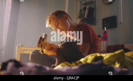 Low angle of glad teenager sitting cross legged on bed near amplifier and playing electric guitar during rehearsal in weekend at home Stock Photo