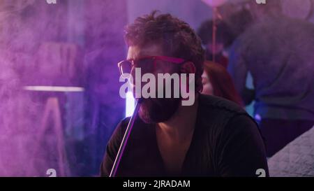 Bearded male in sunglasses exhaling hookah smoke while chilling under neon illumination during party at home Stock Photo