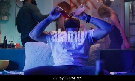 Bearded male drinking alcohol and dancing wildly on couch while having fun during home party with friends at night Stock Photo