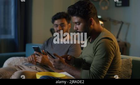 Bearded Indian guy showing smartphone and discussing bets with friends while sitting on sofa and watching sports match on TV in evening at home Stock Photo