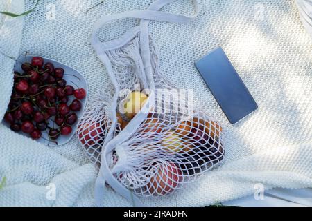 A mesh string bag with fruit, a plate of cherries and a phone on a white background. Picnic in nature in the park. Stock Photo