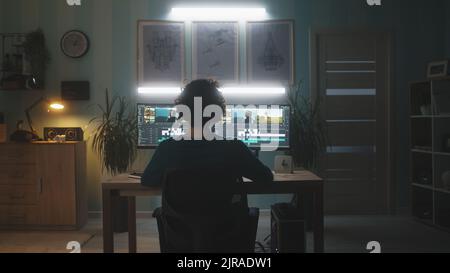 Anonymous male freelancer sitting at desk and editing video on computer in dark home office in evening Stock Photo