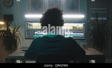 Anonymous man with curly hair editing video on computer while working on remote project in dim workplace at home Stock Photo