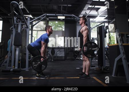 Profile full length shot of twin brothers bodybuilder doing barbell deadlift workout, standing face to face at gym Stock Photo