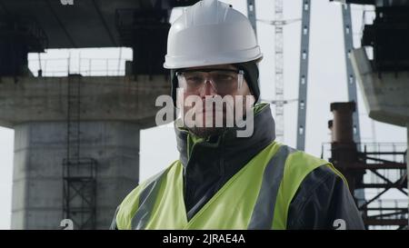 Man in protective goggles and hardhat looking at camera with serious face against concrete bridge supports on construction site Stock Photo