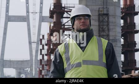 Man in hardhat and goggles looking away while standing under unfinished bridge during work on construction site Stock Photo