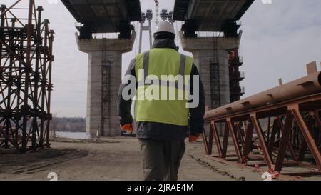 Man in uniform and hardhat walking on dusty path under unfinished bridge during work on construction site Stock Photo