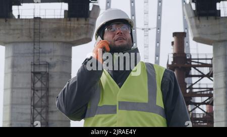 Man in hardhat and goggles inspecting unfinished bridge and talking on smartphone with boss during work on construction site Stock Photo
